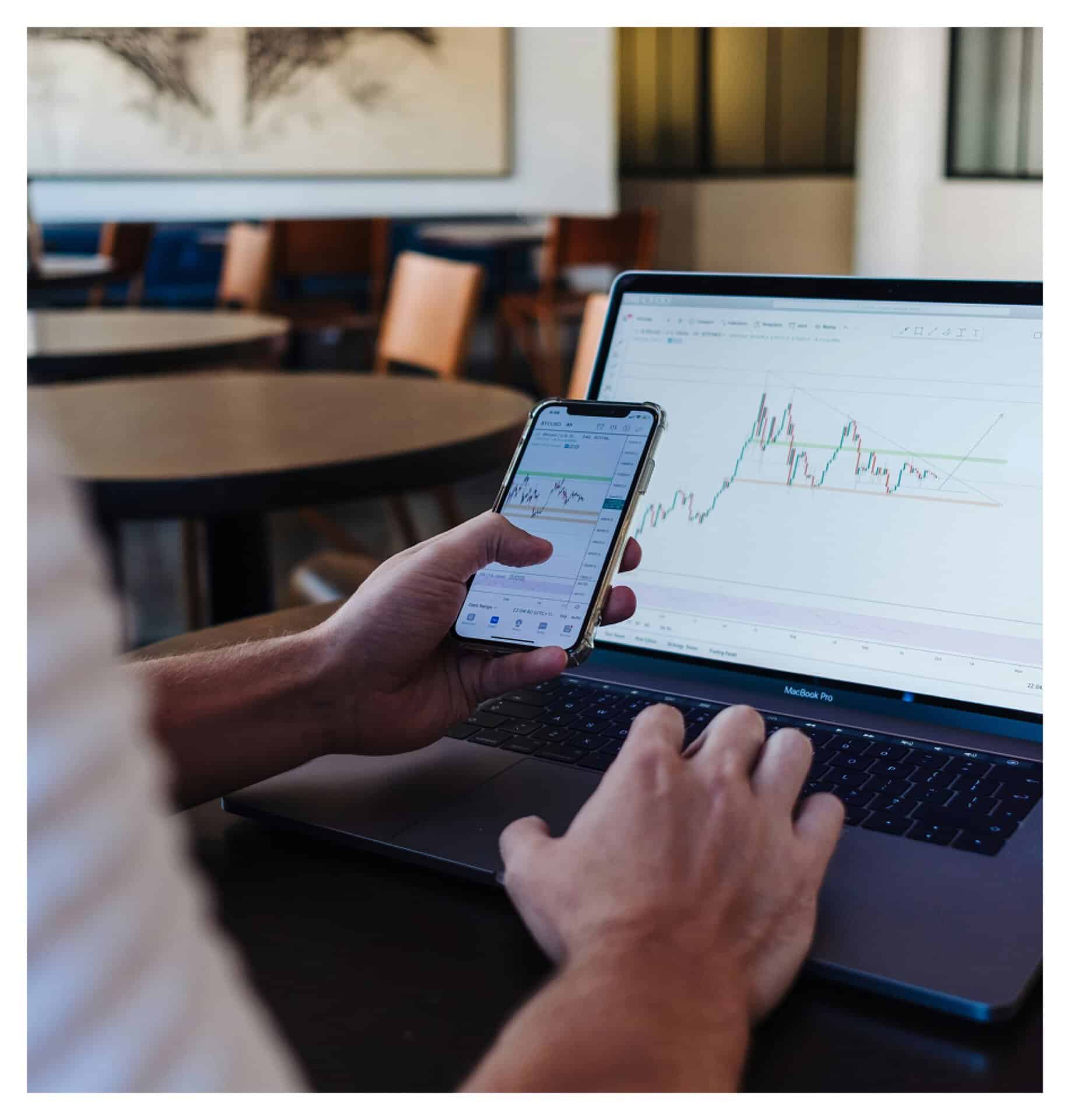 Person reviewing stock market charts on laptop and smartphone in a cafe setting.